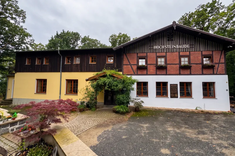 Hochsteinbaude zepředu: hrázděný štít s nápisem Hochsteinbaude, vlevo tmavá dřevěná přístavba, před ní vchod pod zeleným listím