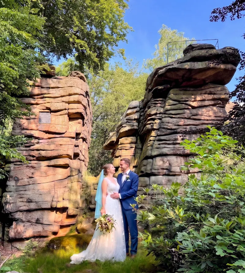 Braut und Bräutigam stehen lachend zwischen zwei hohen Granitfelsen am Hochstein, sie im weißen Kleid mit Brautstrauß und türkisfarbenem Schleier, er im blauen Anzug